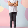 Young girl wearing a white t-shirt and dark leggings standing against a colorful abstract background.